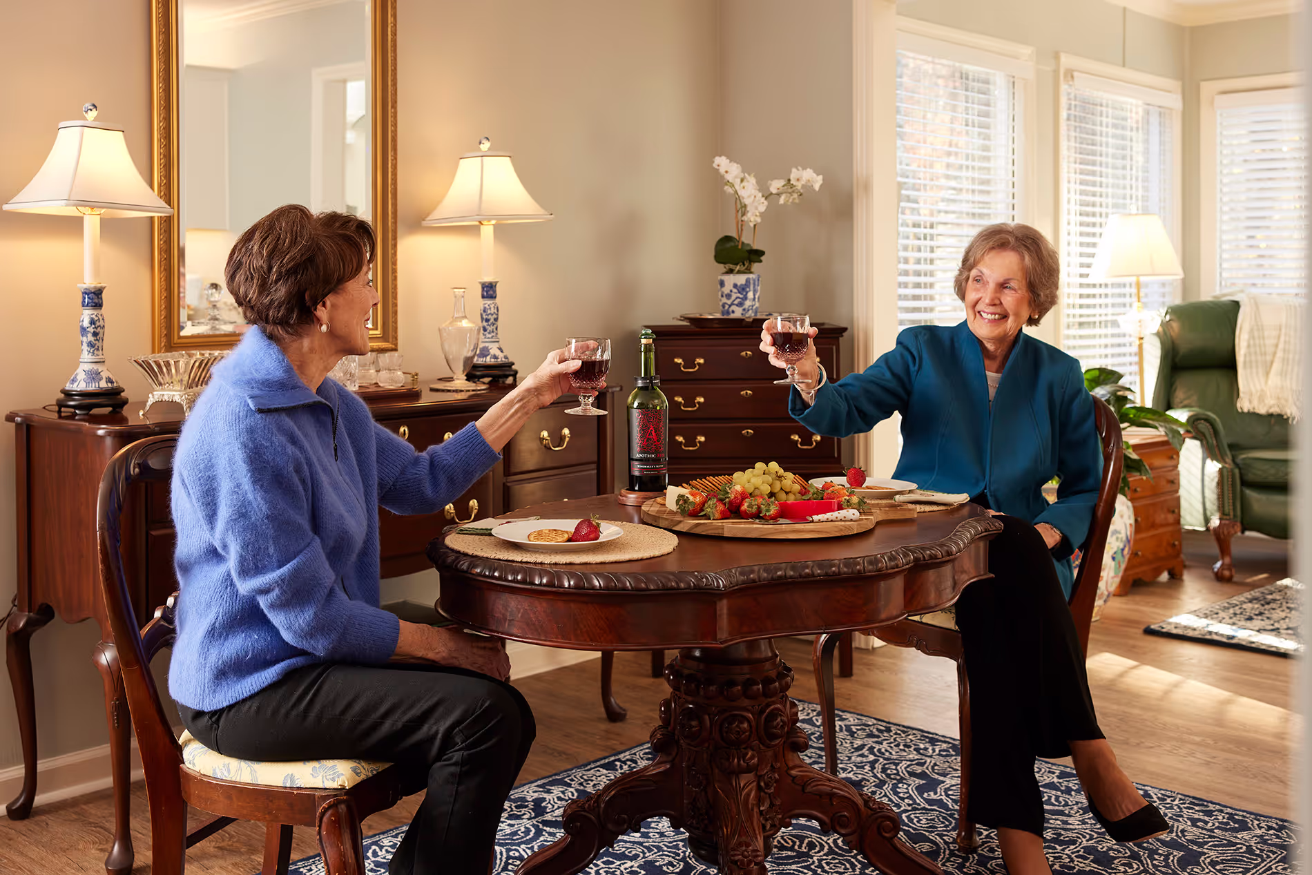 Two women sit at a table, smiling and toasting with glasses of red wine.