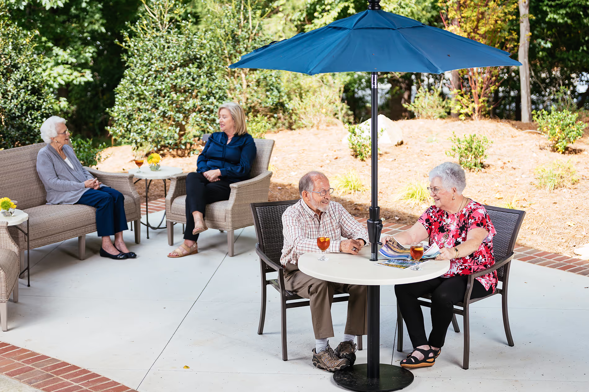 An elderly couple enjoys drinks at an outdoor table under a blue umbrella, while others sit nearby.
