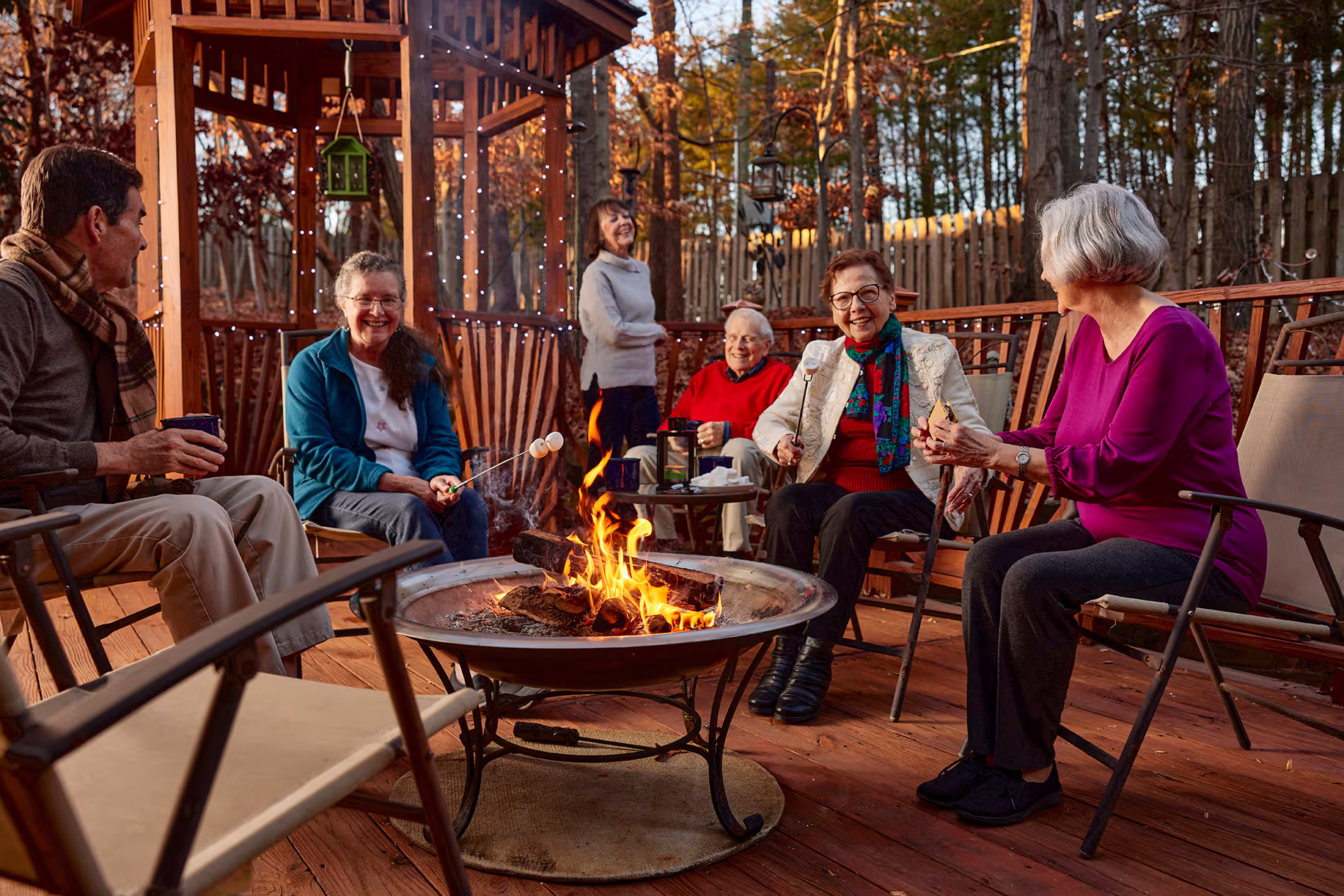 A group of older adults sit around a fire pit, talking and roasting marshmallows outdoors.