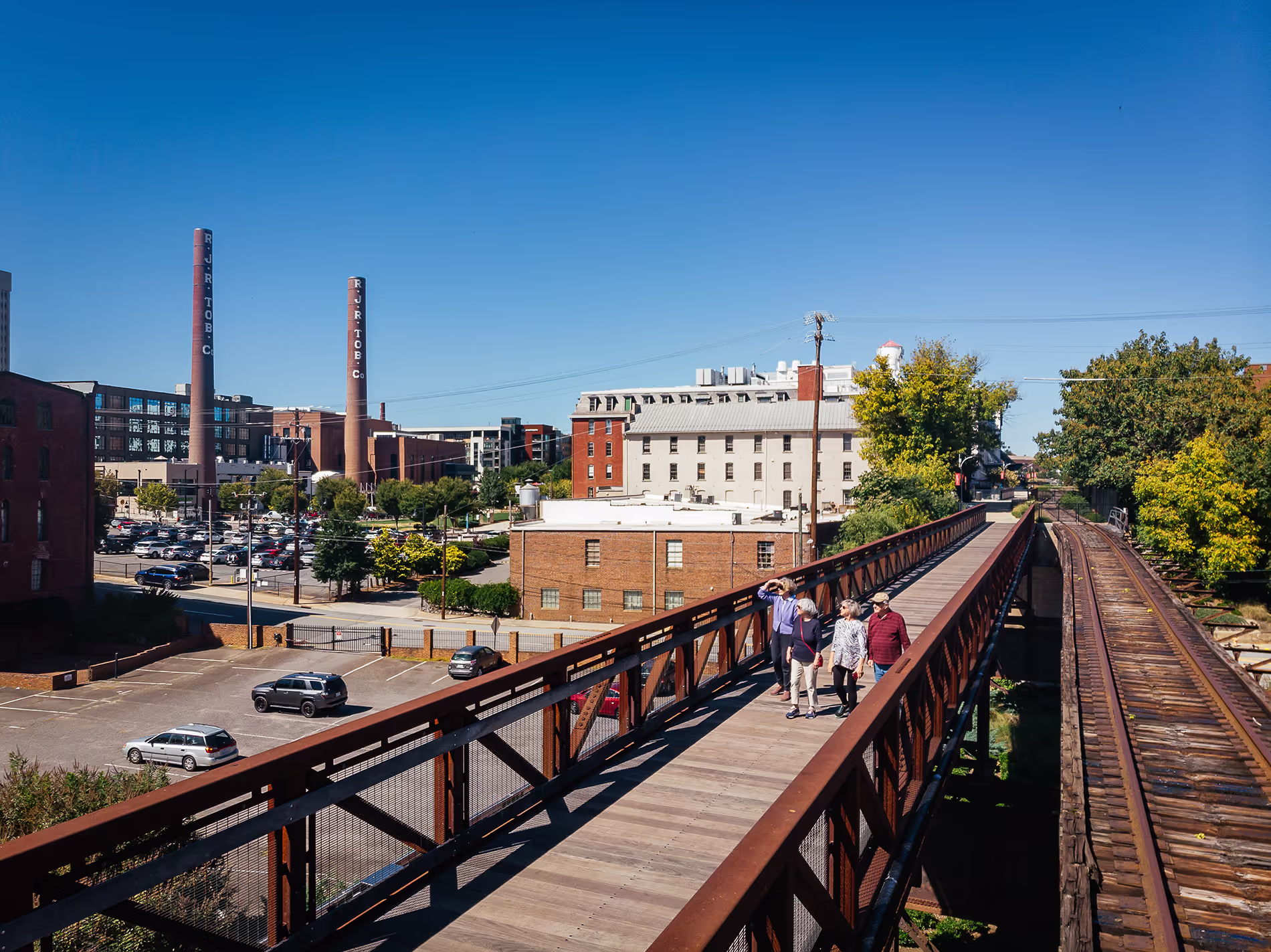 Four people walk on a wooden bridge toward brick buildings and two tall smokestacks.