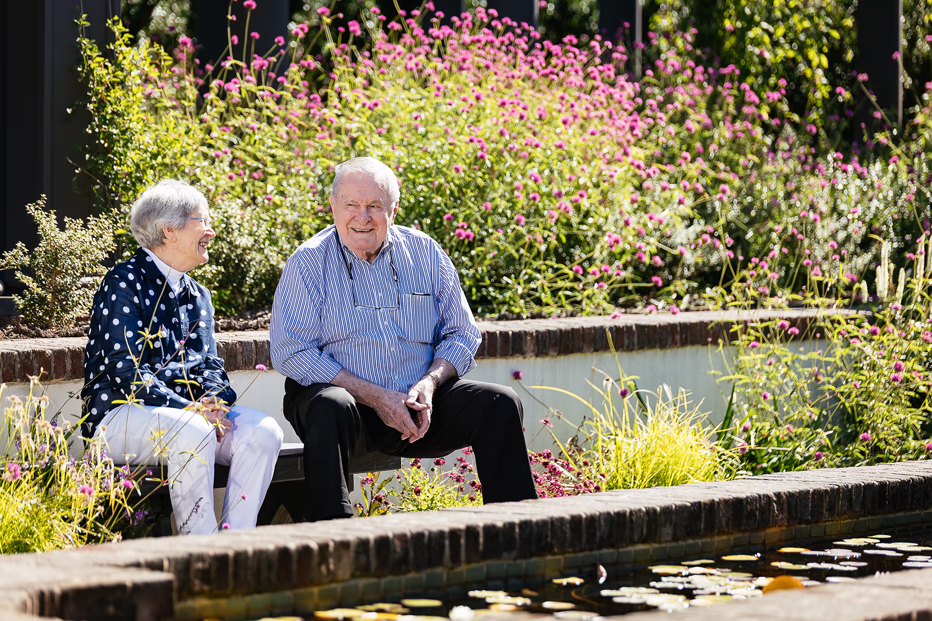 An elderly couple sits and smiles on a bench in a garden with blooming flowers.