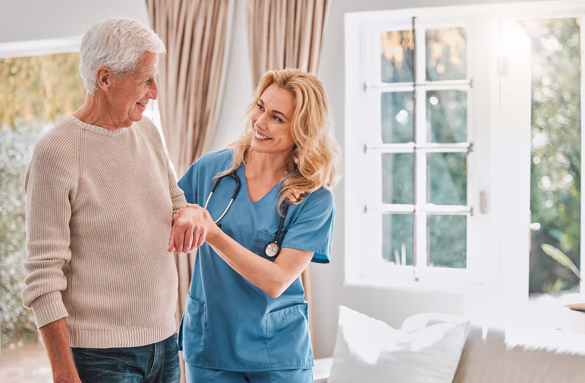 A nurse in blue scrubs assists an elderly man in a beige sweater inside a room.