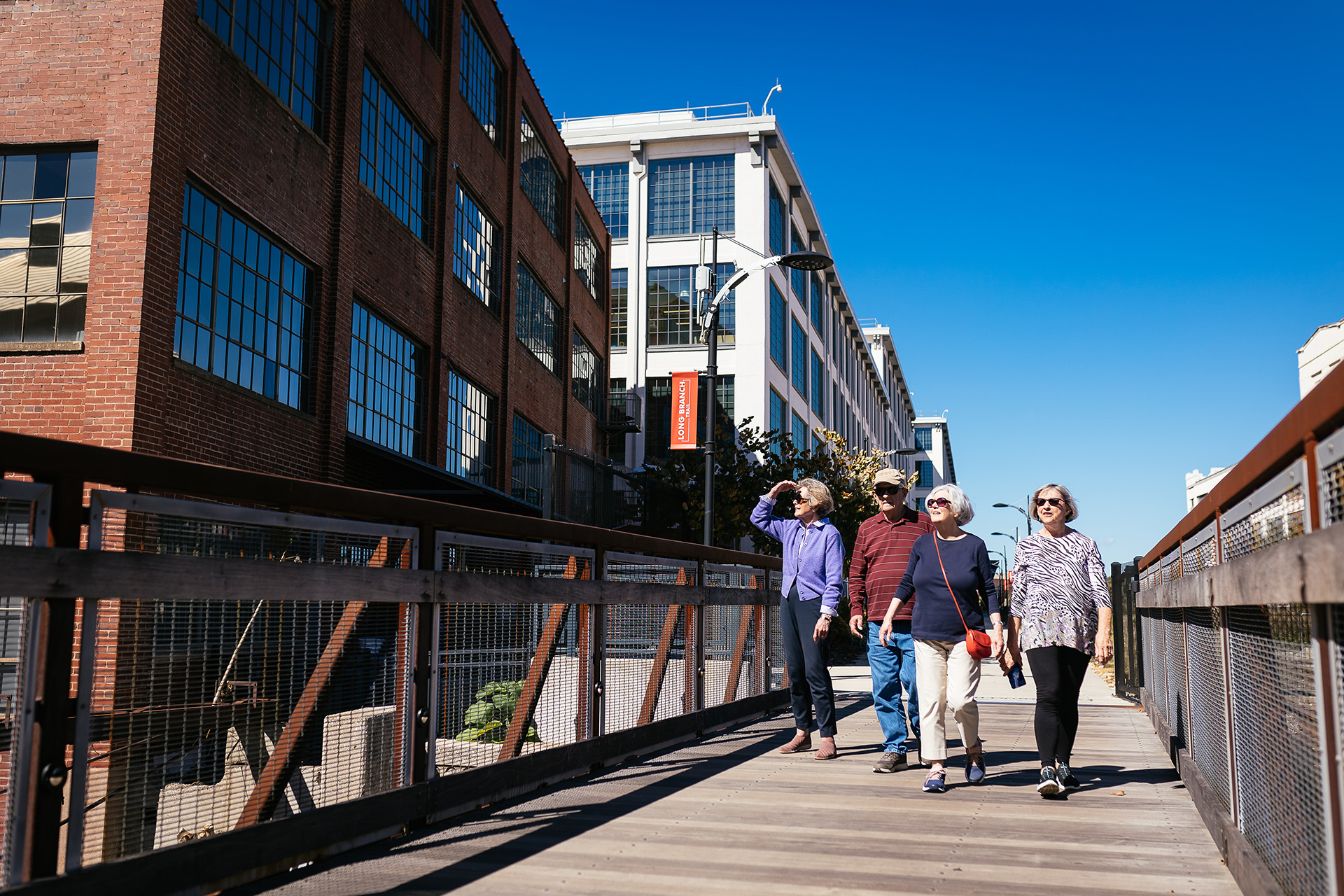 Four people walk across a wooden bridge near tall buildings on a sunny day.
