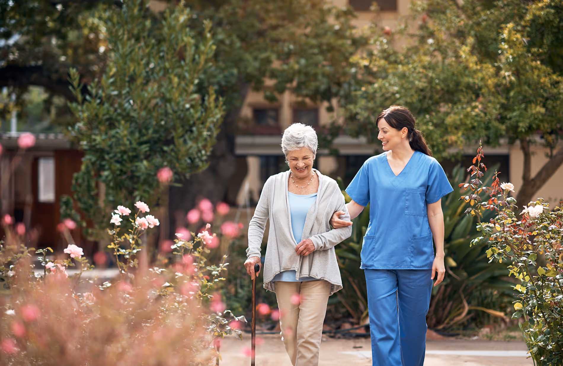 A nurse assists an elderly woman with a cane as they walk through a garden.