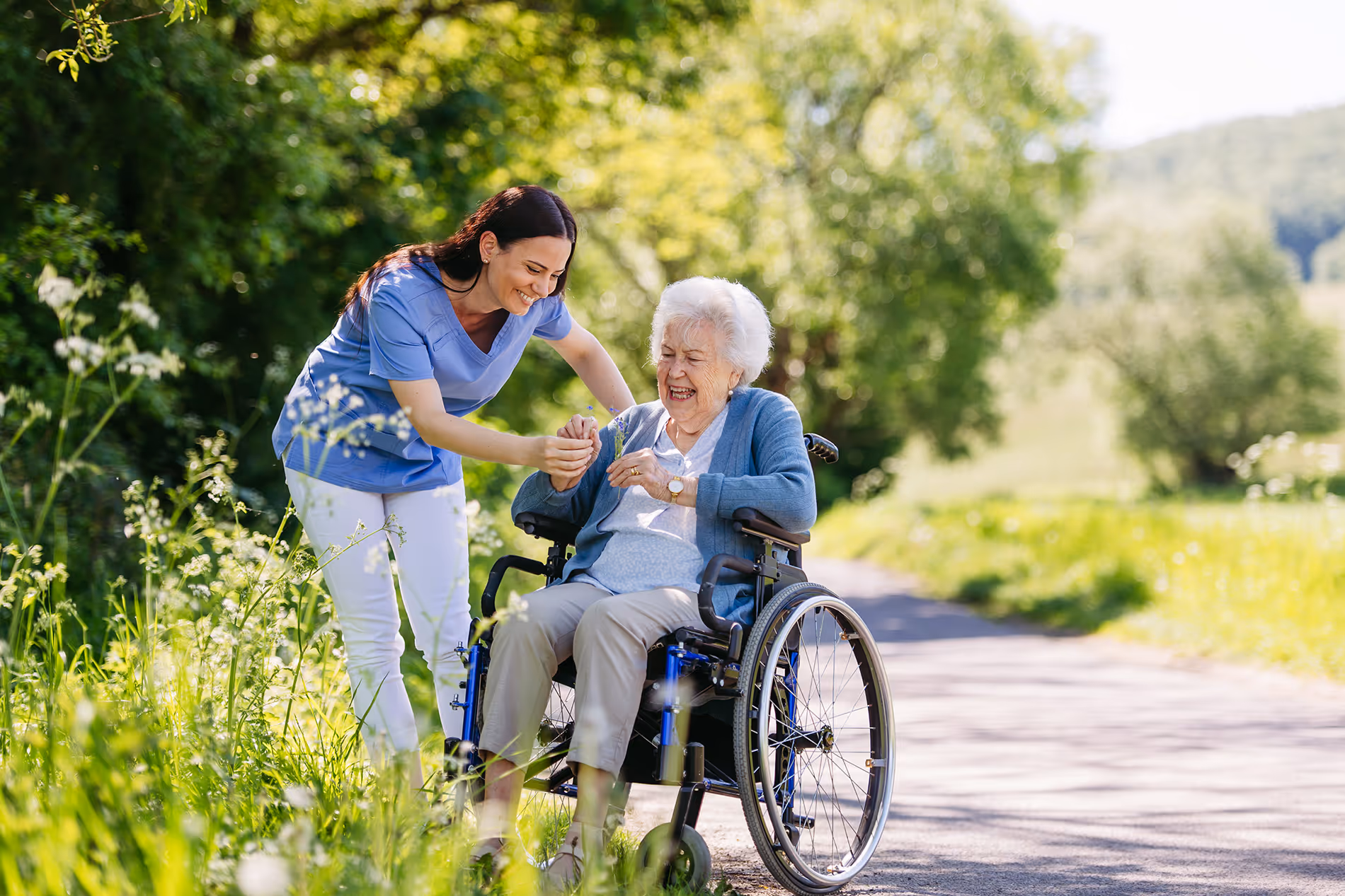 A woman assists an elderly woman in a wheelchair on a sunny path outdoors.