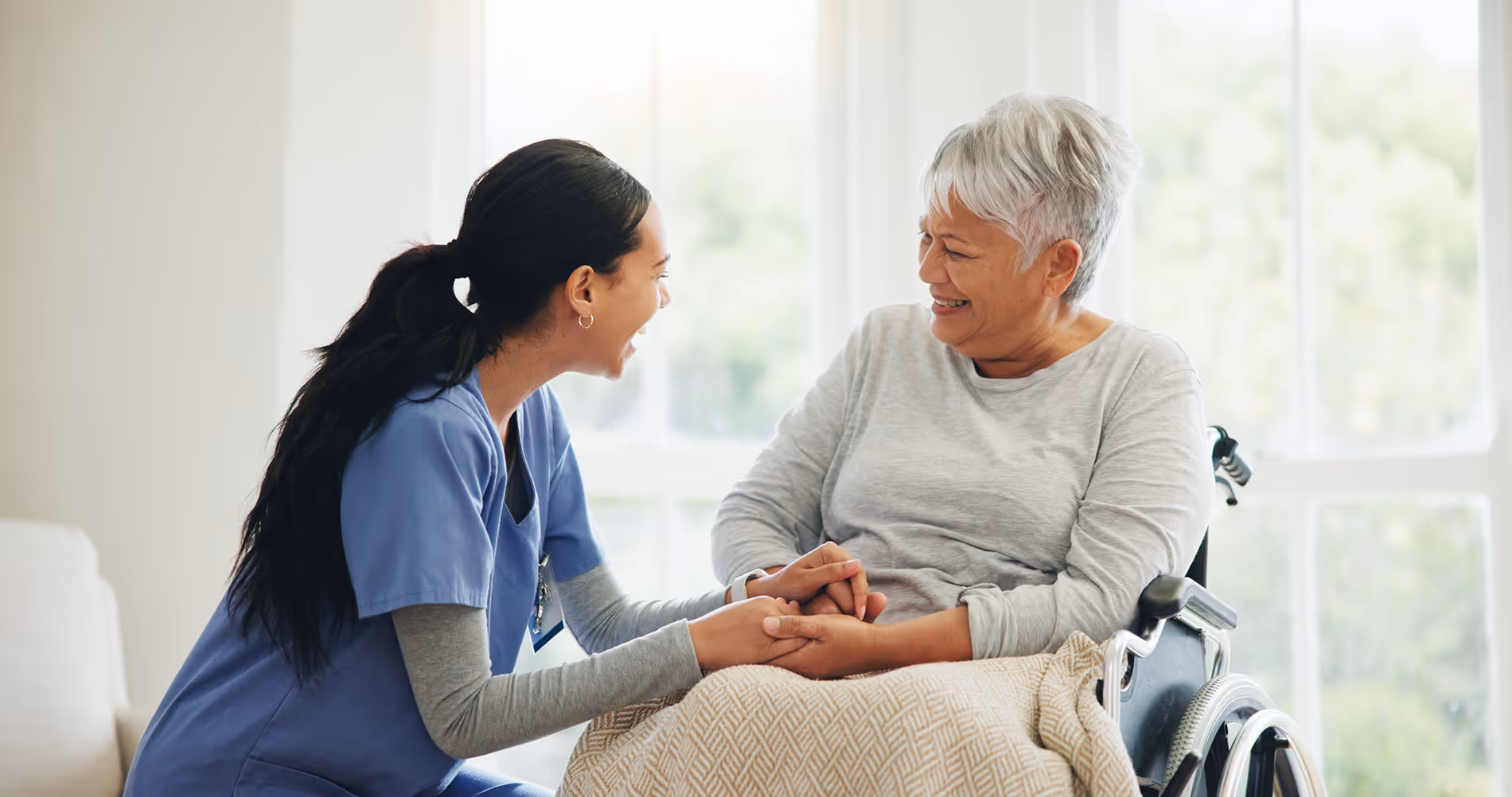 A nurse smiles and holds hands with an older woman sitting in a wheelchair.