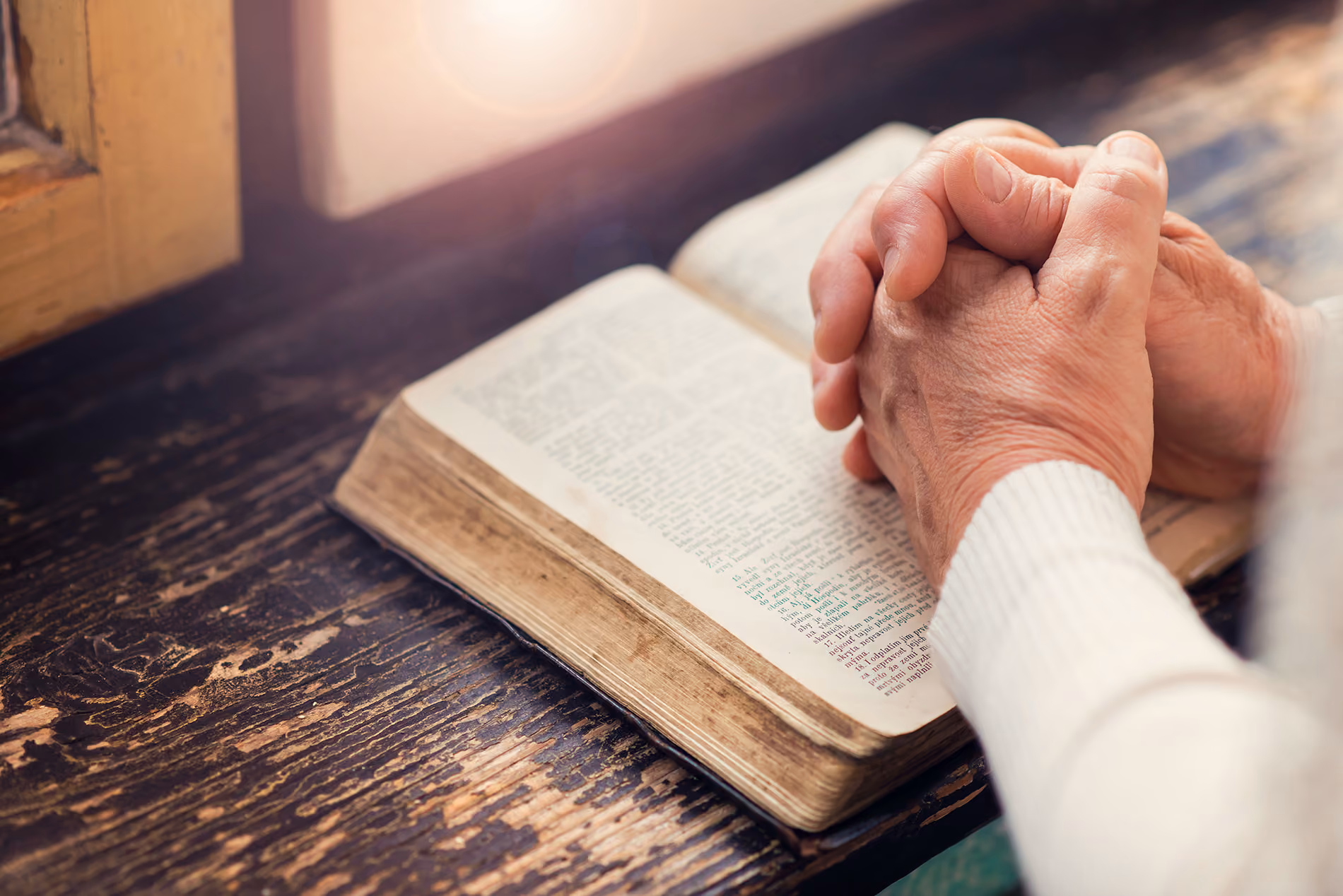 Hands clasped in prayer resting on an open, worn book on a wooden surface near a window with light.