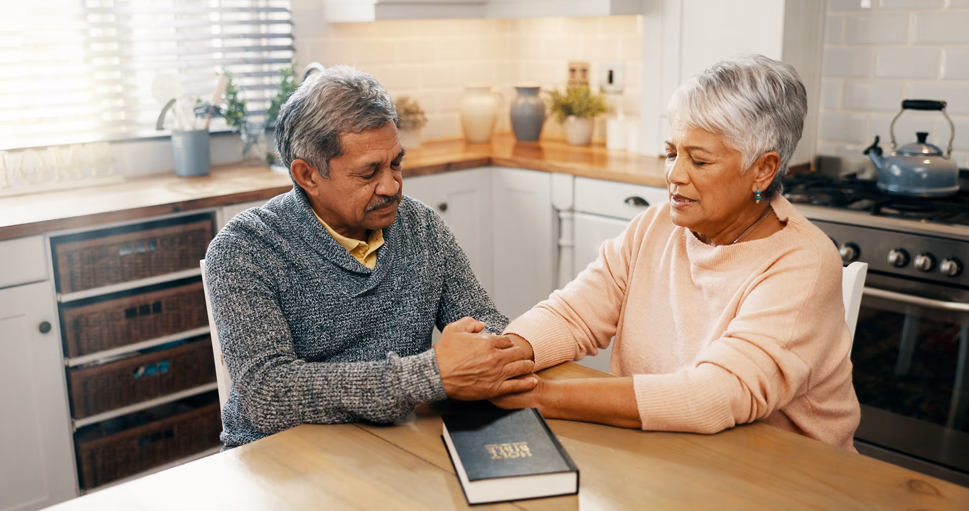 Older couple sitting at kitchen table holding hands, with a Holy Bible resting on the table.