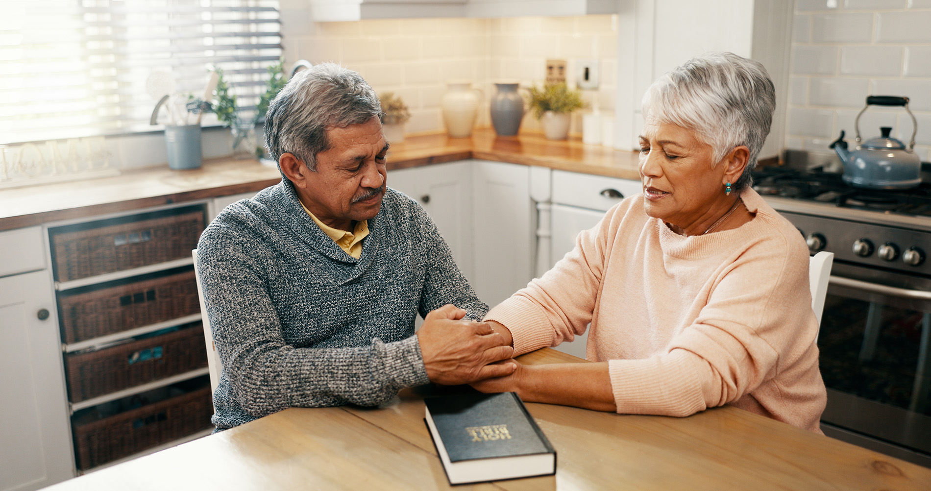 Older couple sitting at kitchen table holding hands, with a Holy Bible resting on the table.