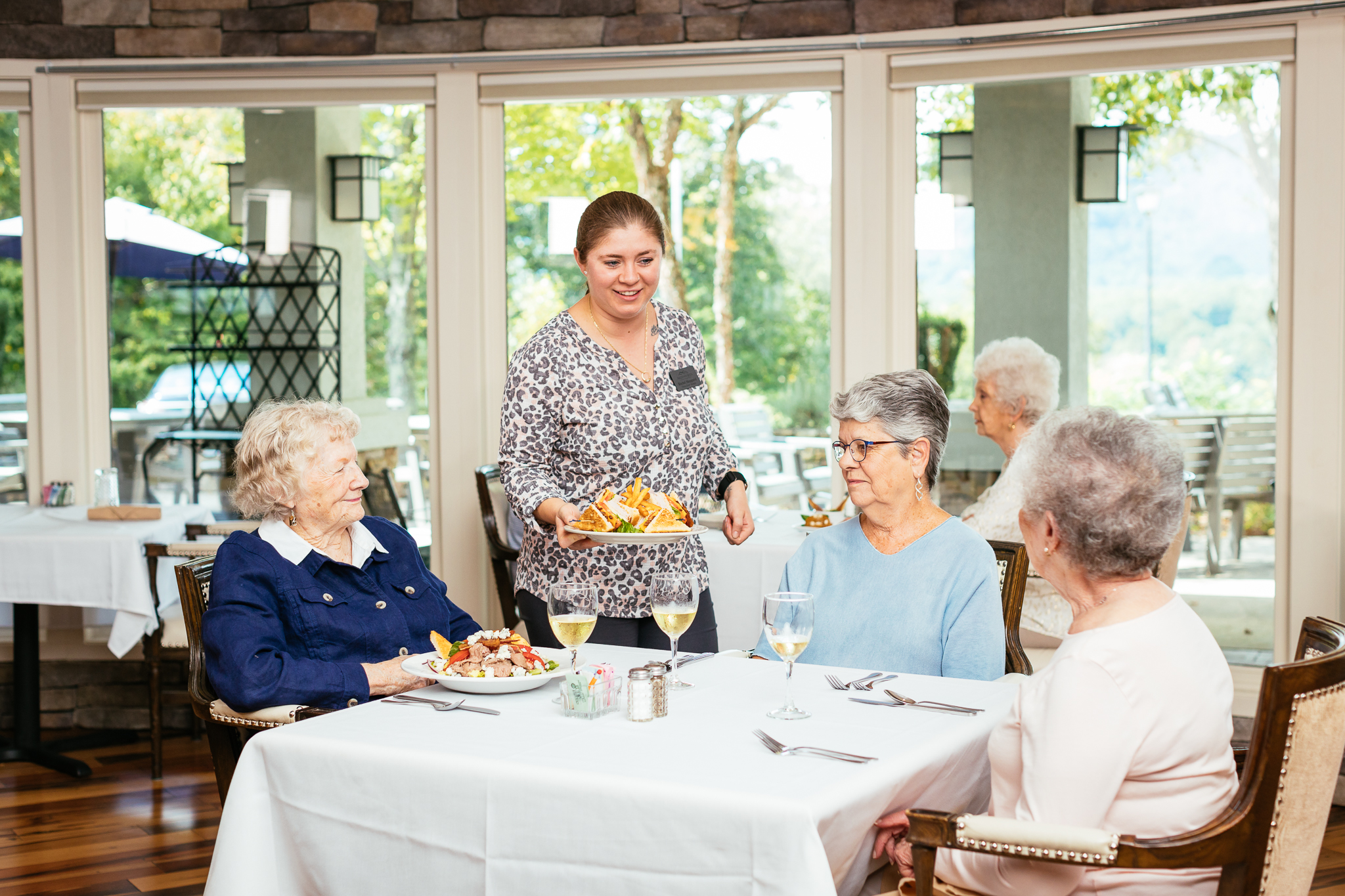 Server placing a plate of sandwich and fries before three elderly women dining at a white-clothed table.