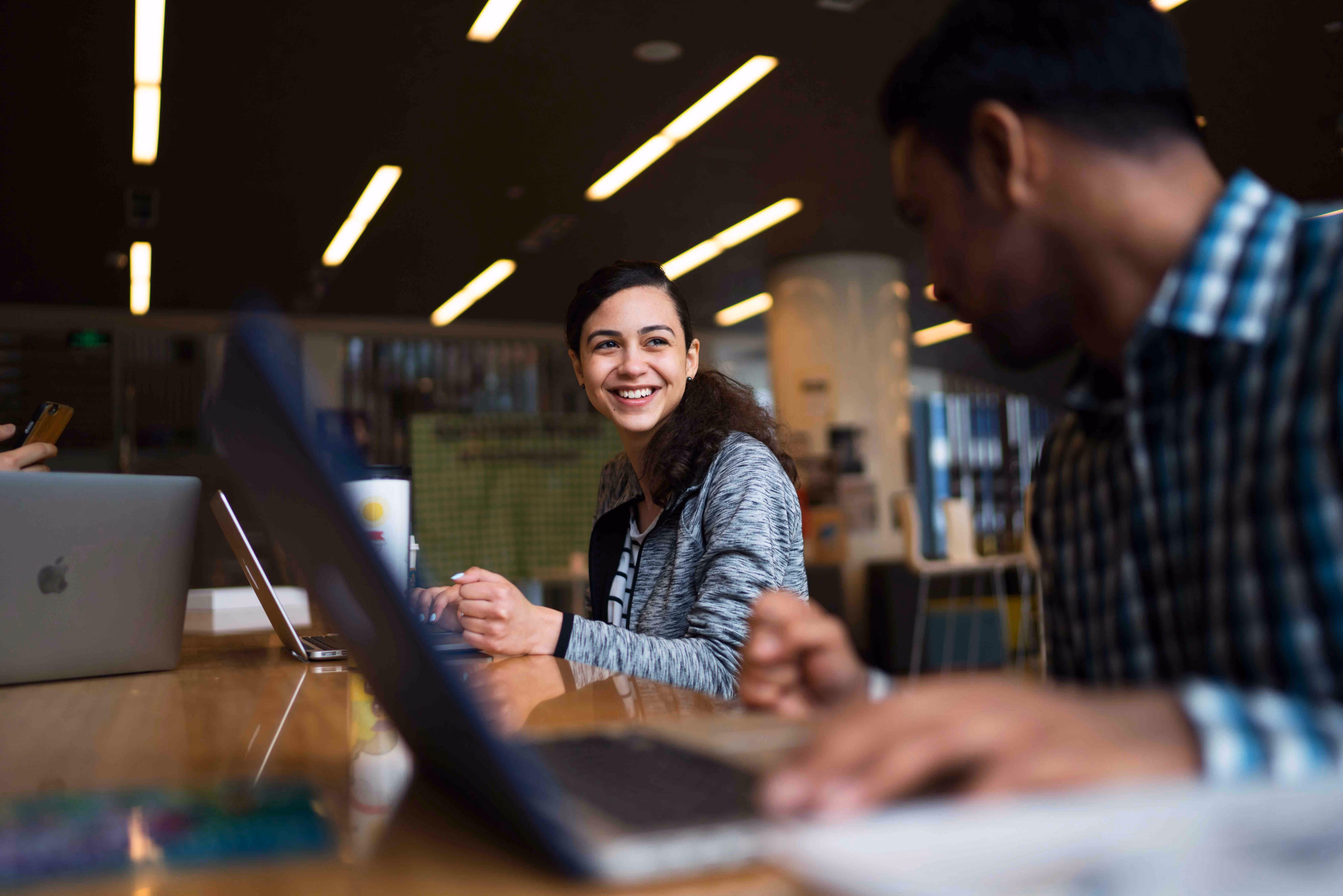 A student smiling and looking at a classmate while working at a laptop in a brightly lit university library or study area.