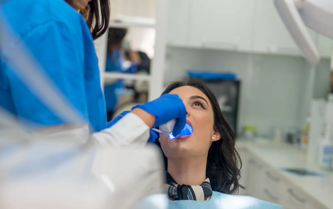 "A female patient undergoing a dental procedure sits reclined in a dental chair as a dentist or dental hygienist, wearing blue gloves and a white coat, uses a curing light device inside her mouth. The blue LED light illuminates the patient’s teeth, likely indicating a bonding, whitening, or composite resin treatment. The clinical setting is clean and modern, with dental tools and equipment visible in the background