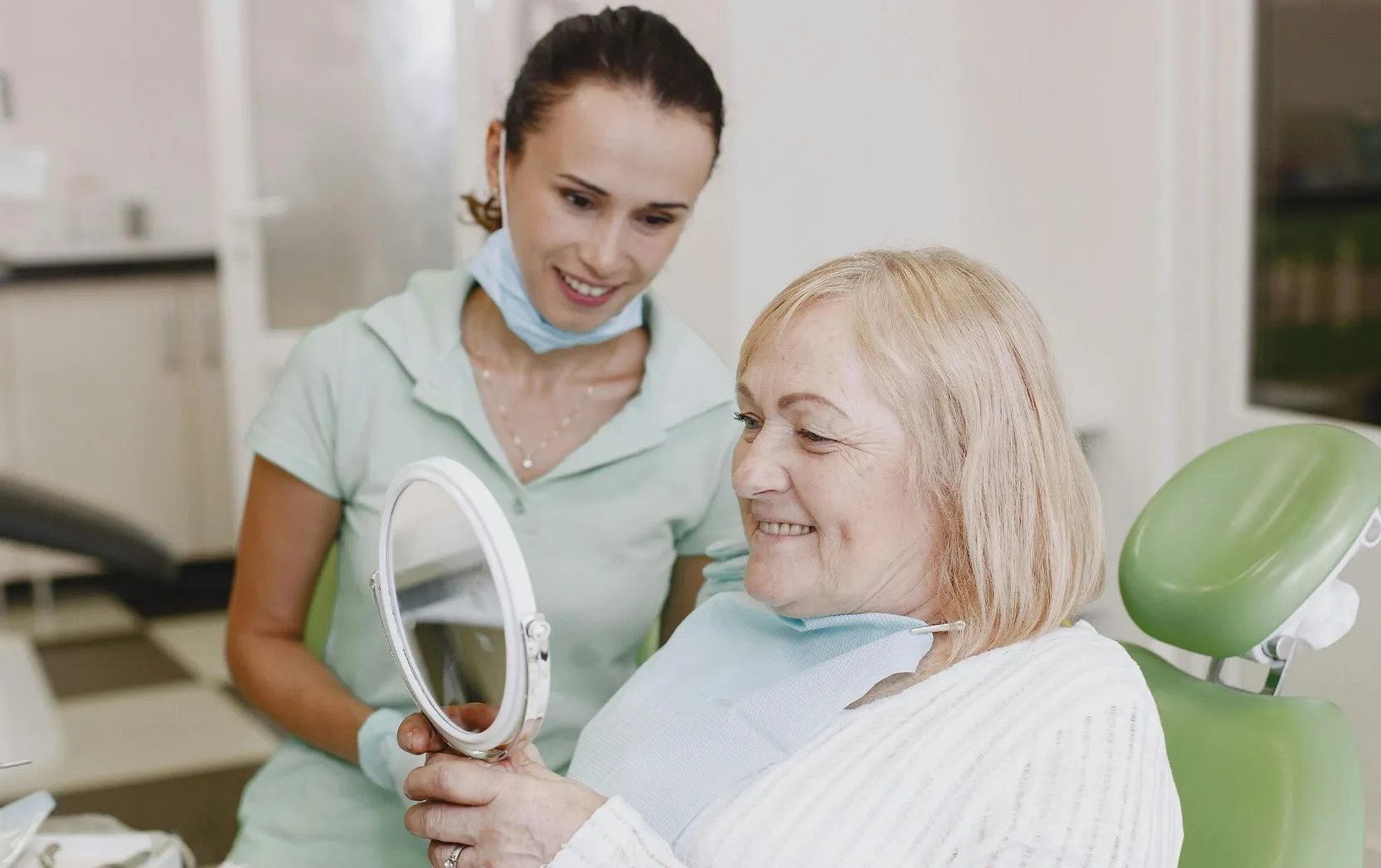 An older female dental patient with blonde hair smiles as she examines her teeth using a handheld mirror while seated in a green dental chair. A female dental professional, dressed in light green scrubs with a face mask lowered under her chin, stands beside her, smiling supportively. The setting is a clean and well-lit dental office, capturing a positive post-treatment moment of satisfaction and care
