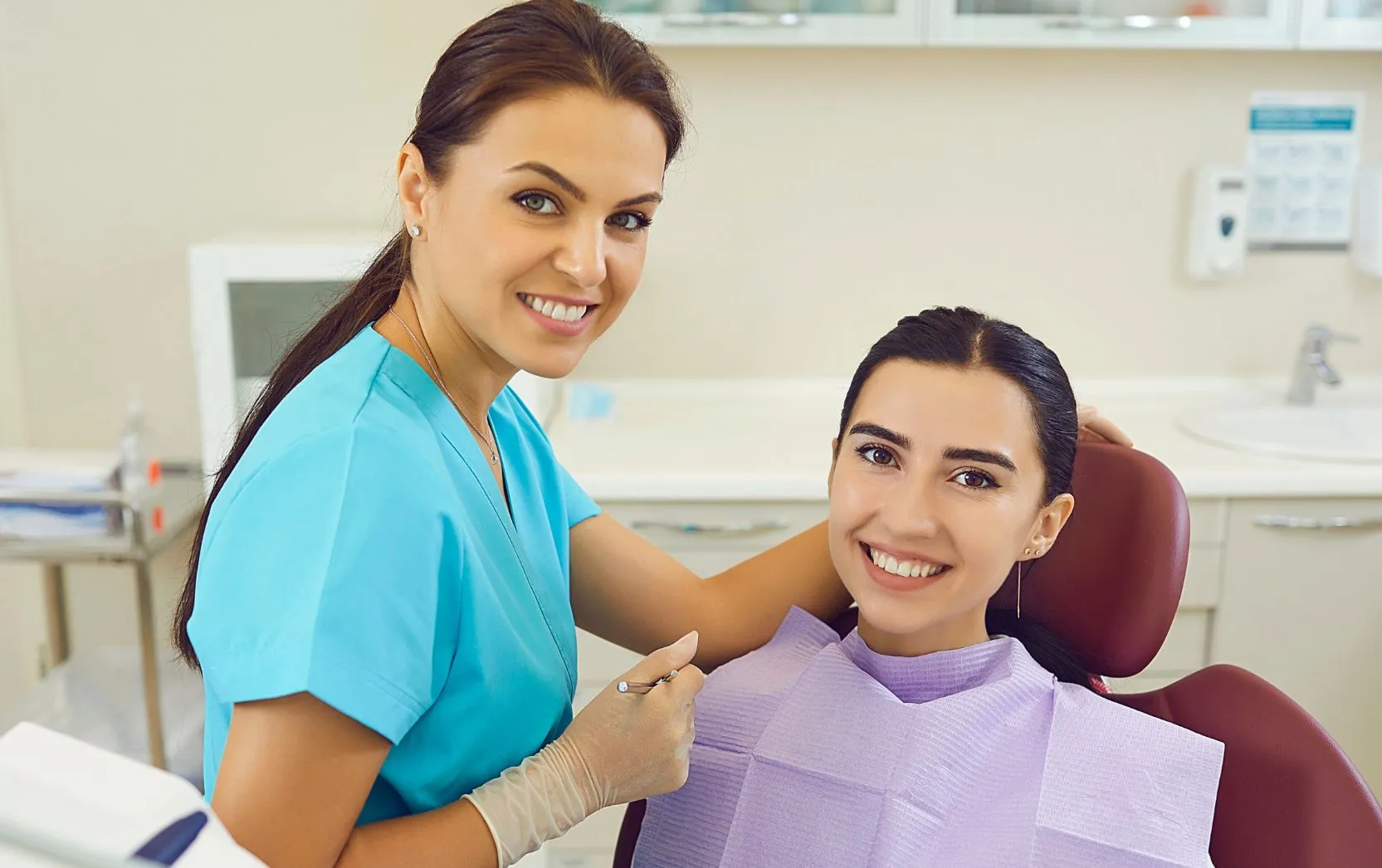 A smiling female dentist wearing turquoise scrubs and latex gloves stands beside a seated female patient in a dental chair. Both women are looking directly at the camera with cheerful expressions. The patient wears a lavender dental bib and rests comfortably in a modern, brightly lit dental clinic, creating a warm and welcoming atmosphere that reflects trust and satisfaction with dental care