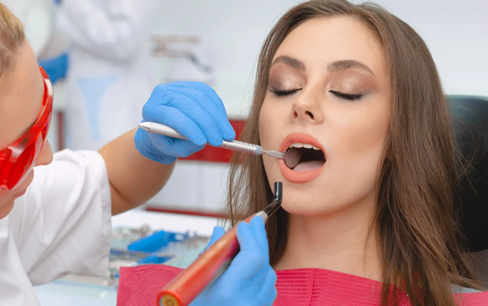A female patient with long brown hair and flawless makeup sits calmly in a dental chair with her eyes closed and mouth open, receiving a dental procedure. A dental professional wearing red protective glasses, a white coat, and blue gloves uses a curing light tool and a dental mirror inside the patient's mouth. The scene suggests a cosmetic or restorative treatment in a modern, well-equipped dental clinic