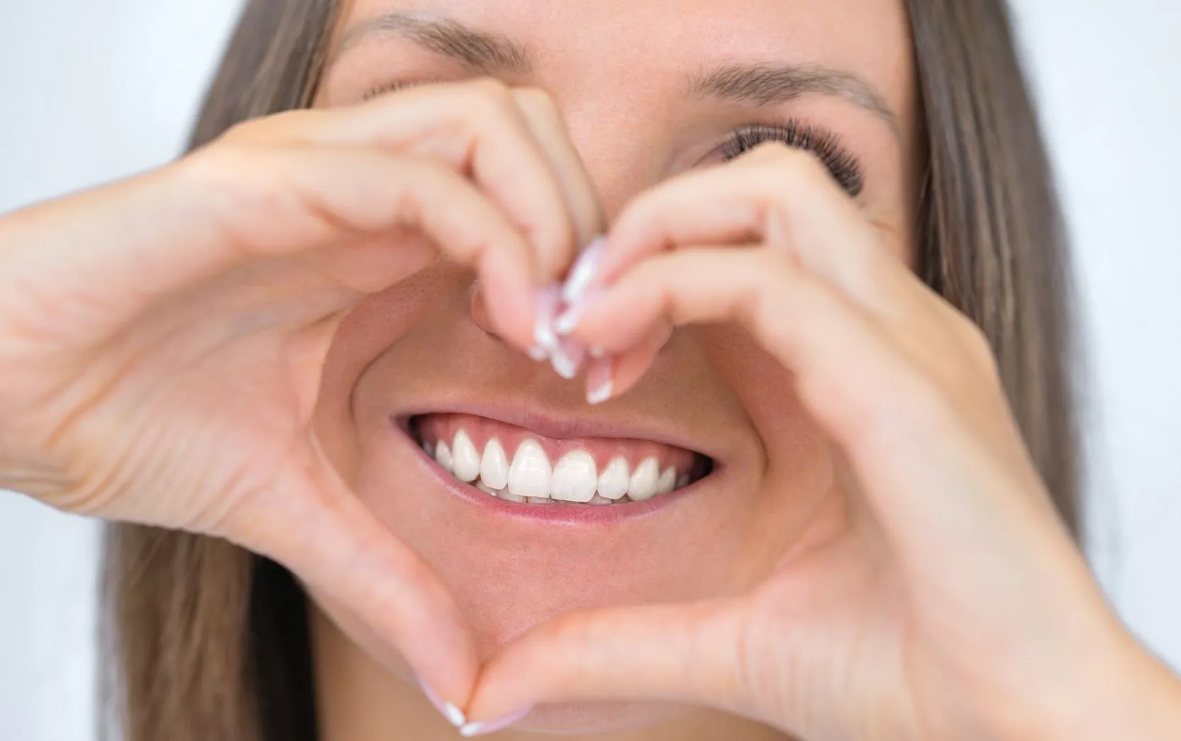 A cheerful woman smiles brightly with flawless white teeth while forming a heart shape with her hands in front of her face, framing her joyful expression and sparkling eyes in a warm and affectionate gesture that radiates positivity and self-confidence