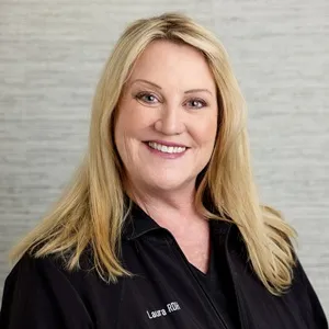 Portrait of Laura, dental hygienist, wearing a black zip-up jacket against a light gray background.