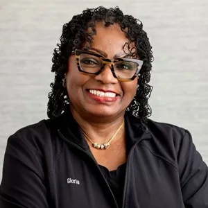 Portrait of Gloria, dental assistant, wearing a black zip-up jacket against a light gray background.