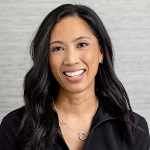 Portrait of Jackie, office manager, wearing a black zip-up jacket against a light gray background.