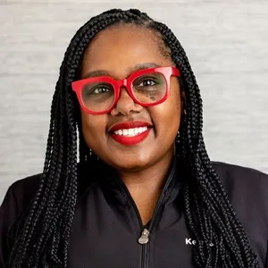 Portrait of Keisha, dental hygienist, wearing red glasses and a black zip-up jacket against a light gray background.