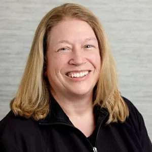 Portrait of Julie, dental hygienist, wearing a black zip-up jacket against a light gray background.