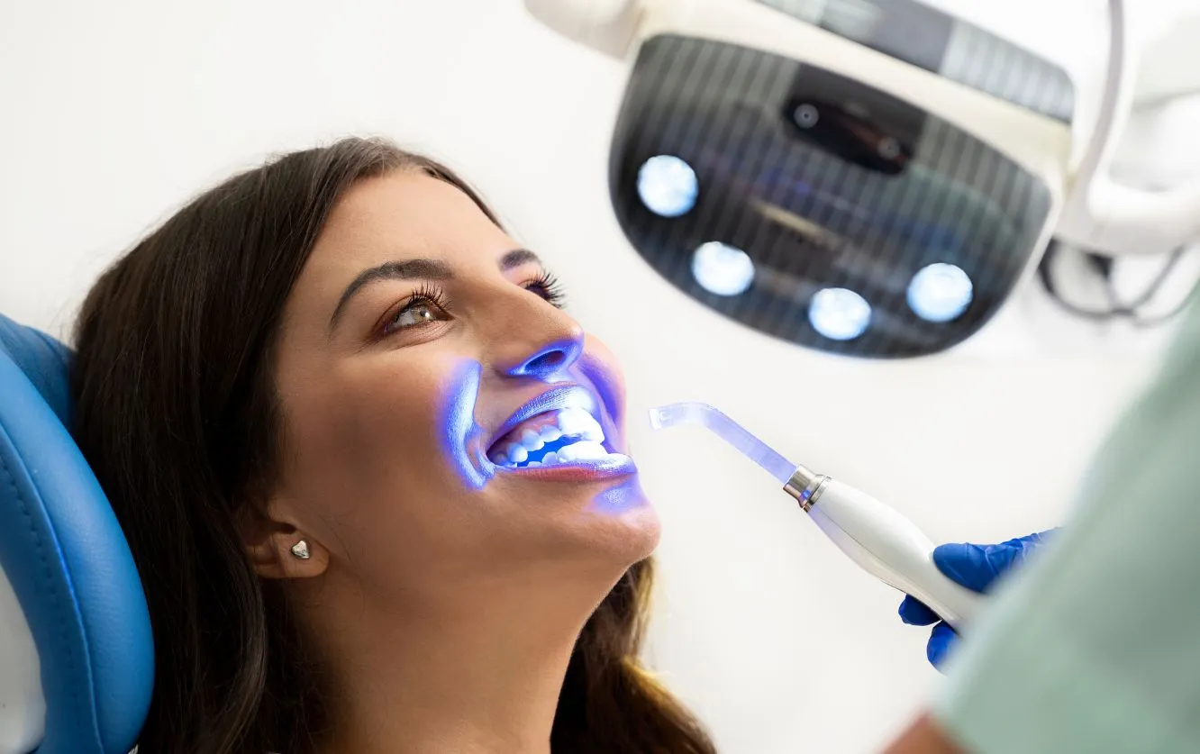 A young woman reclines in a dental chair with her head tilted back and mouth open in a relaxed smile as a dental professional uses a curing light tool to apply focused blue light, illuminating her teeth and cheeks while an overhead surgical lamp casts additional glow, creating a high-tech, clinical atmosphere that captures the precision and care of modern cosmetic dental treatment