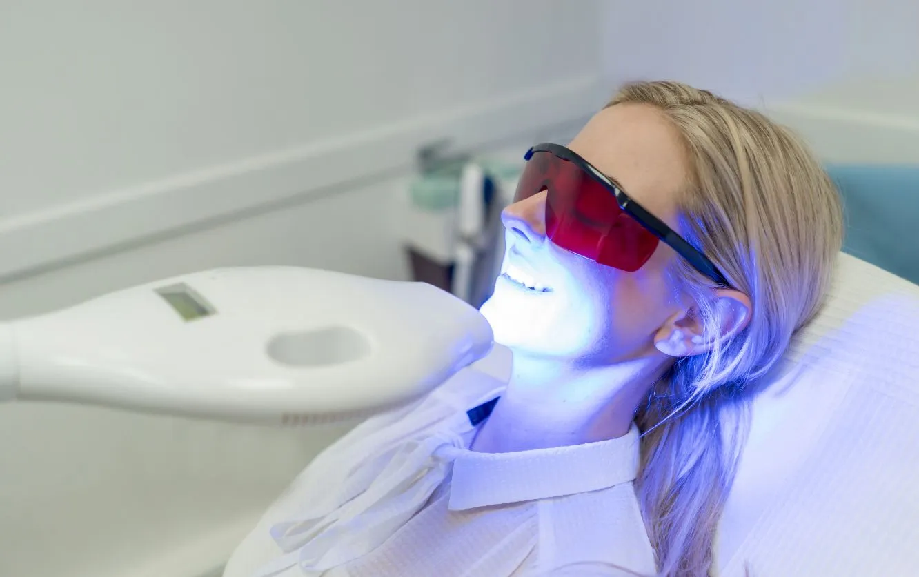 A woman reclines comfortably in a modern dental clinic wearing protective red-tinted glasses while a bright blue LED light from a professional whitening device is directed at her teeth, illuminating her face and the surrounding area as she undergoes an advanced in-office teeth whitening procedure designed to enhance her smile by lifting deep-set stains from the enamel