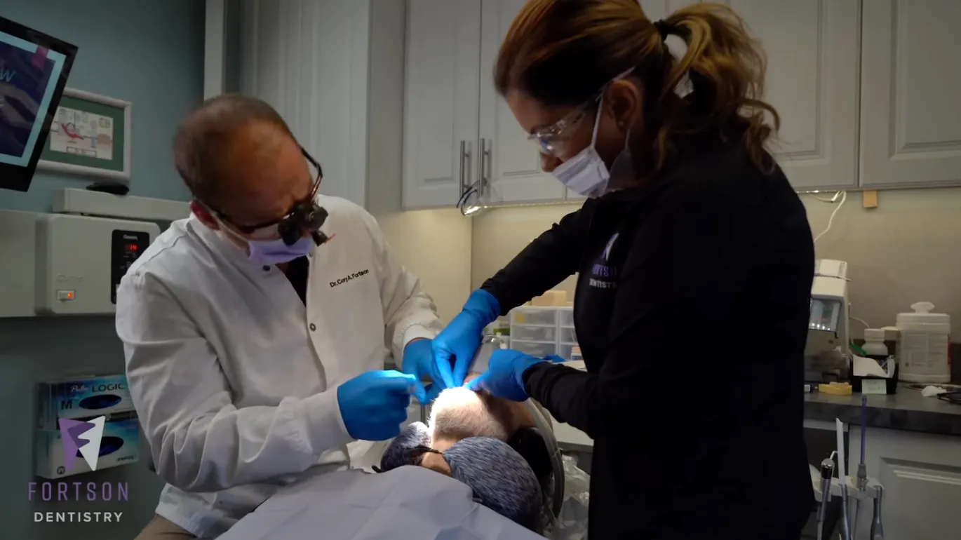 Two dental professionals performing a procedure on a patient lying in the dental chair, with both wearing gloves, face masks, and working under overhead lighting in a modern operatory