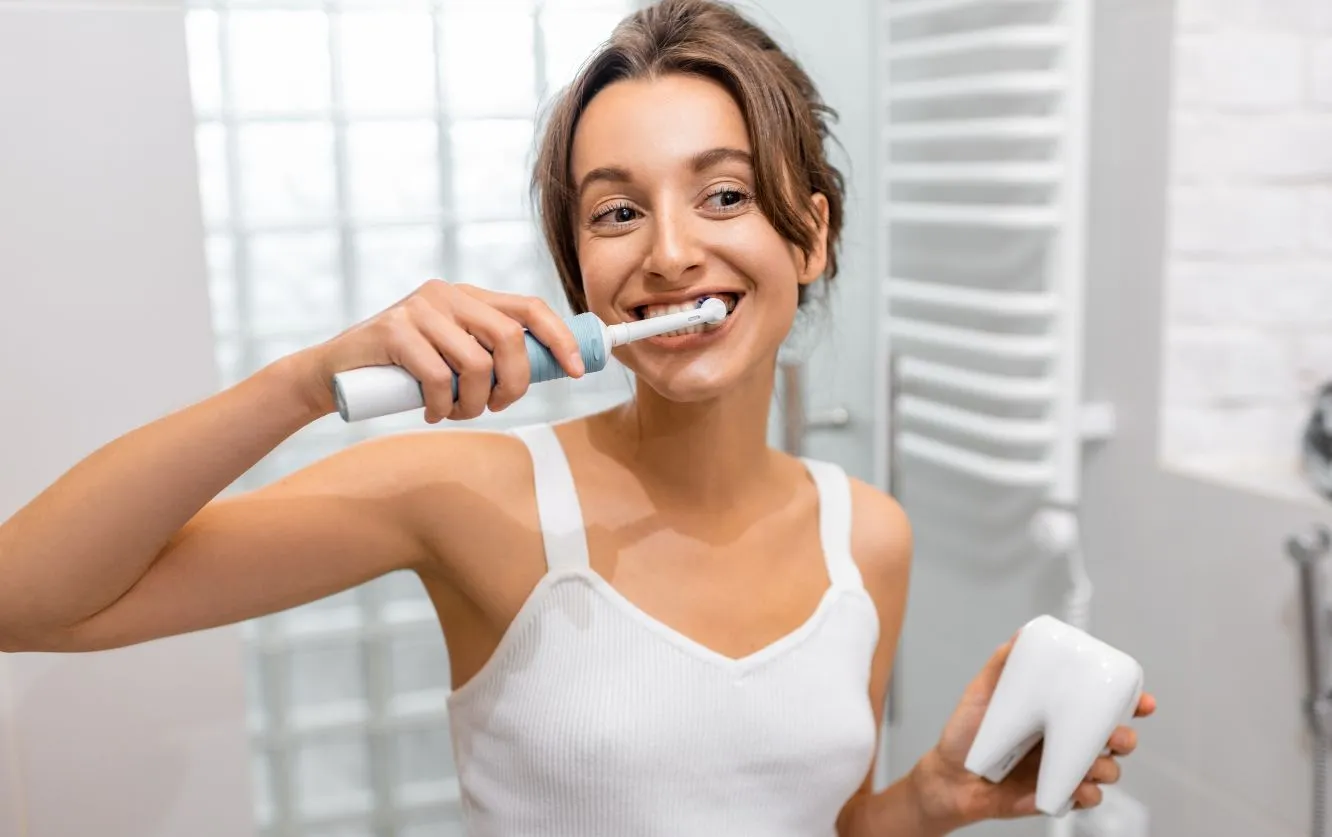 Smiling woman brushing her teeth with a white manual toothbrush in a well-lit bathroom, demonstrating proper hygiene