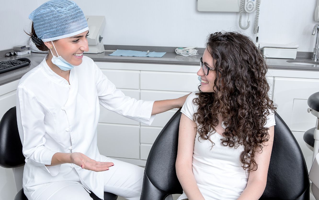 Dentist in surgical scrubs greeting a female patient seated in the operatory before beginning a dental examination