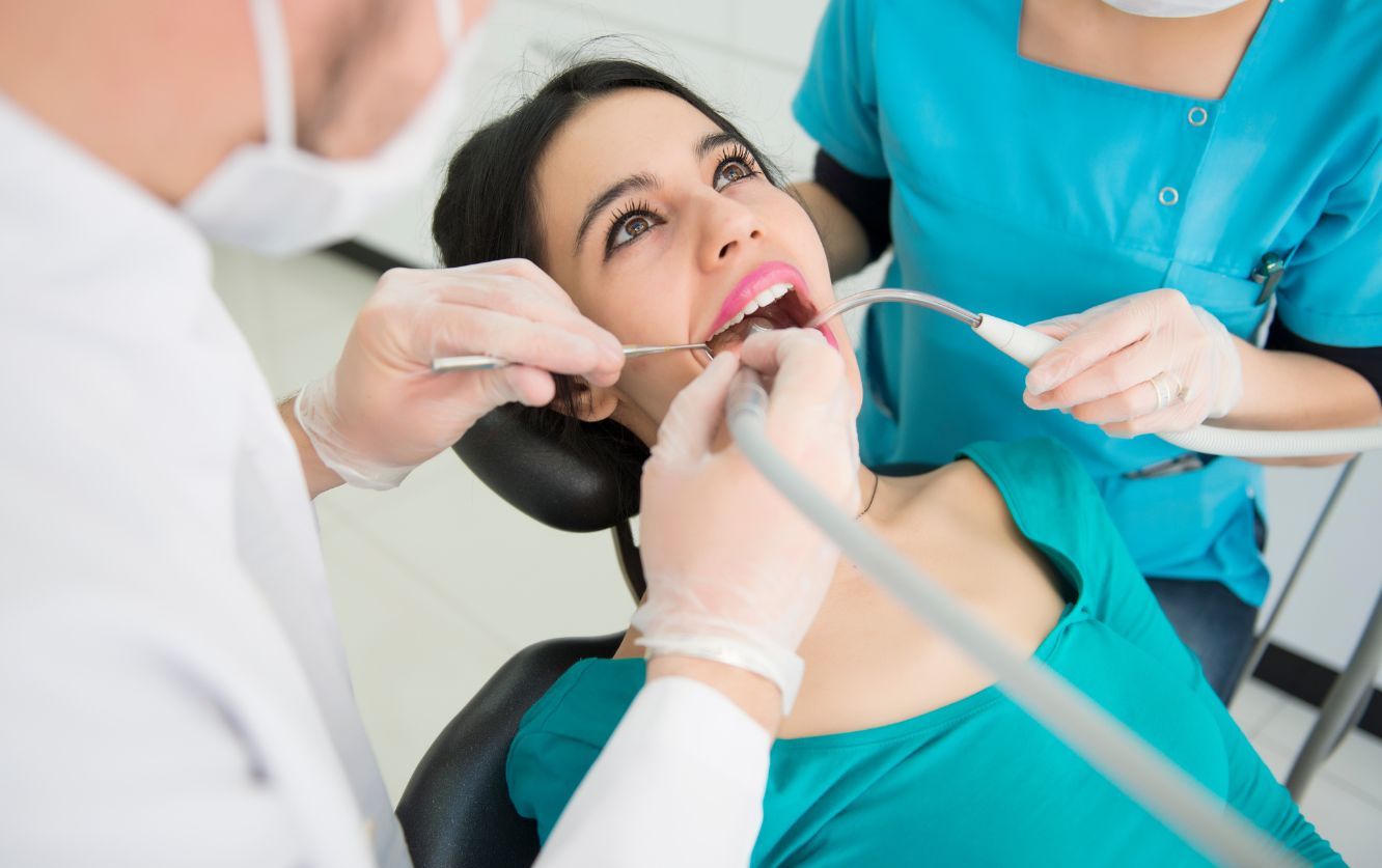 Female patient in dental chair receiving treatment while dentist and assistant examine her teeth using a mirror and scaler