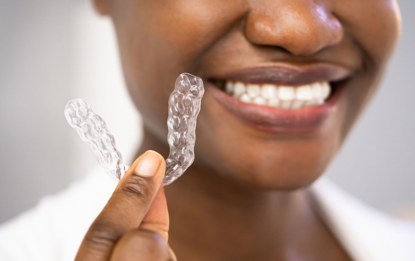 Close-up of a woman holding a clear aligner between her fingers and smiling, demonstrating Invisalign orthodontic treatment