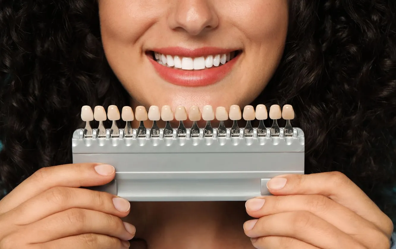 Smiling patient holding a shade guide up to her teeth as a cosmetic dentist compares color swatches to determine the best match for veneers or whitening treatment