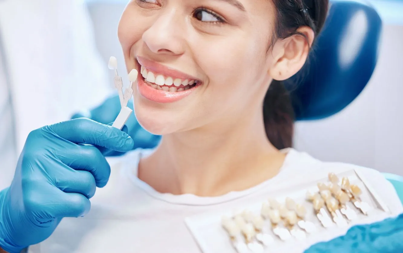 Cheerful young woman pointing to her bright white teeth while a gloved dental assistant holds a whitening shade guide to highlight results after a professional teeth whitening session