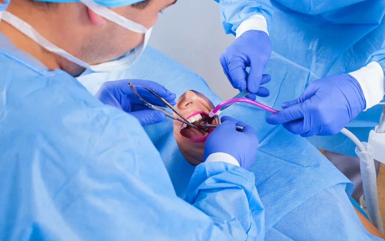 Dental surgeon and assistant wearing blue surgical gowns, gloves, and protective masks during a sterile in-office oral surgery procedure under bright LED lighting