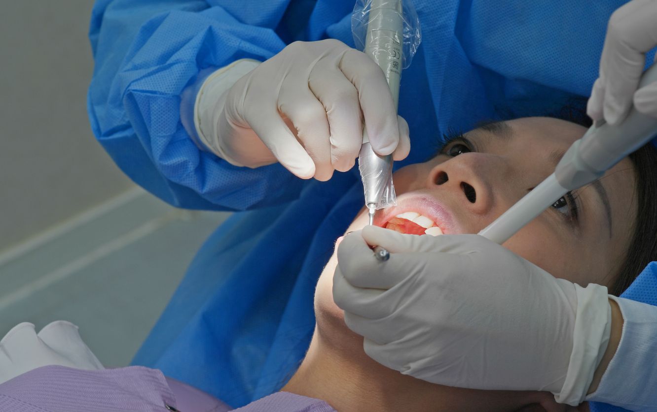Close-up of a dental assistant wearing sterile blue scrubs and gloves preparing a metal syringe filled with anesthetic solution and organizing stainless steel dental instruments on a surgical tray before a procedure