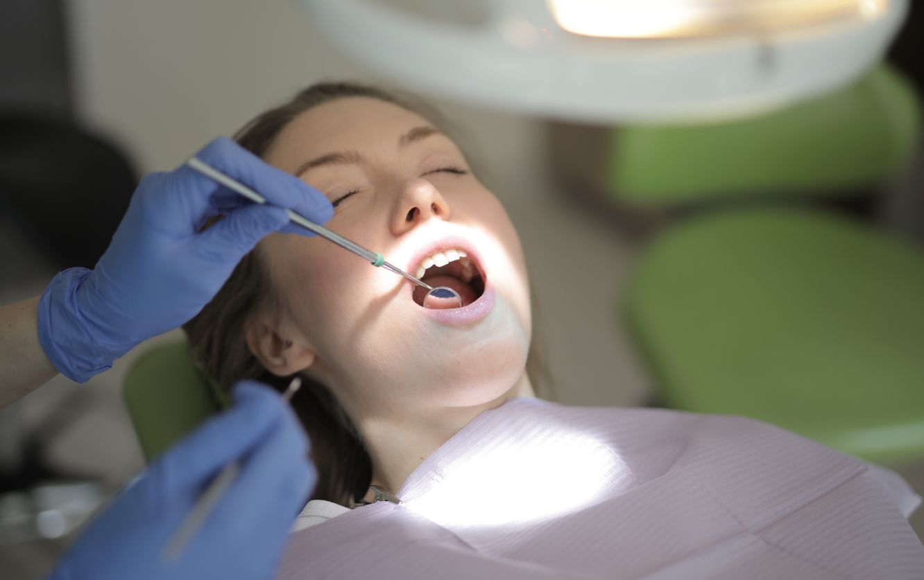 Young child reclining in a modern dental chair, smiling with mouth wide open under an overhead exam light while wearing protective orange safety glasses during a pediatric dental cleaning session