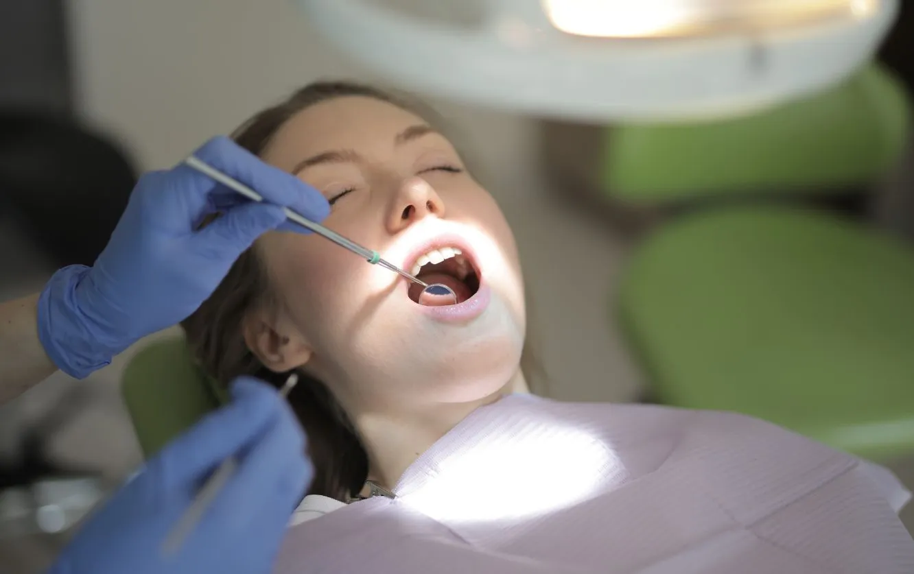 Young child reclining in a modern dental chair, smiling with mouth wide open under an overhead exam light while wearing protective orange safety glasses during a pediatric dental cleaning session