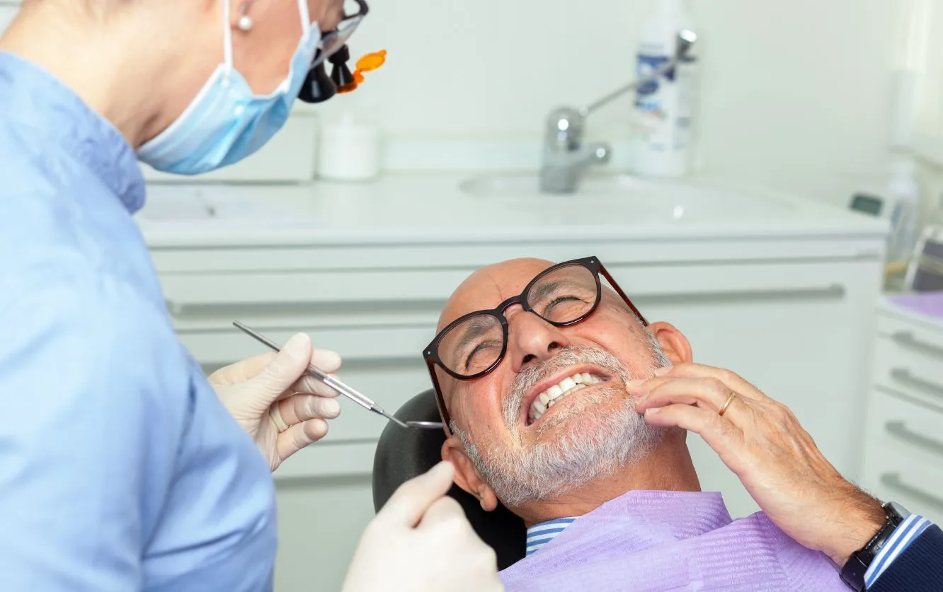 Senior male patient wearing glasses receiving a dental exam from a masked hygienist using a dental mirror and scaler at a modern dental clinic, showing slight discomfort during the cleaning procedure