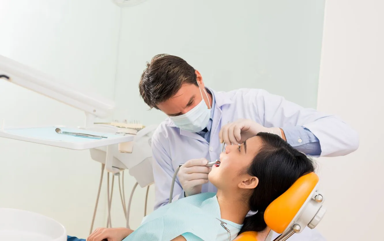 Male dentist wearing a face mask performing a dental examination on a female patient reclining in a yellow dental chair using a mirror and scaler tool inside a clean and modern dental clinic environment