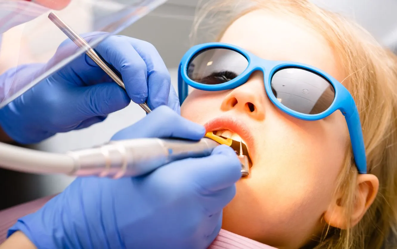 Young child wearing blue sunglasses receiving a professional dental cleaning from a pediatric dentist using a scaler and suction tool, with gloved hands visible, demonstrating gentle and kid-friendly dental care in a bright clinical setting