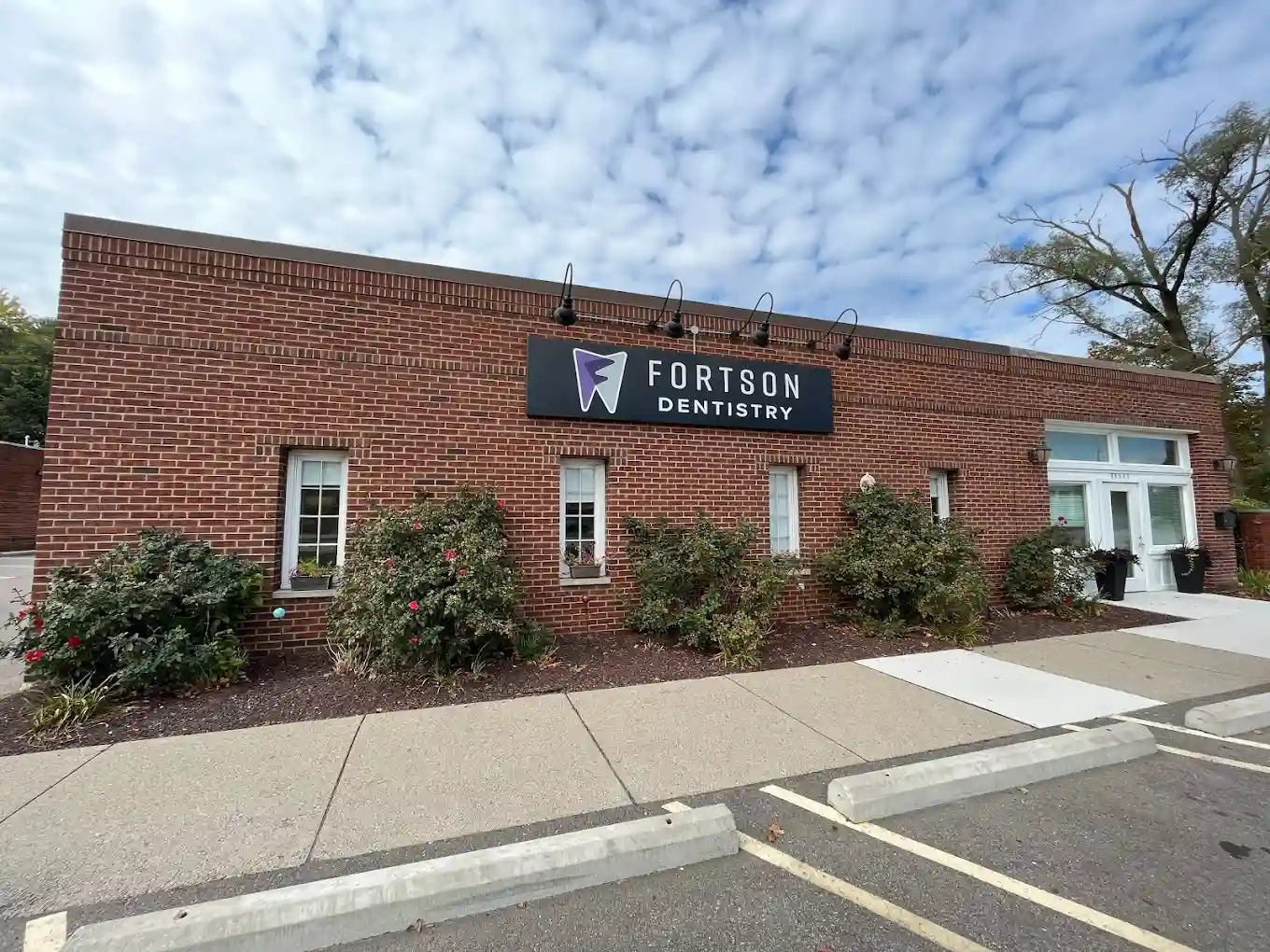 Exterior view of Fortson Dentistry in Lathrup Village, Michigan, showing a modern brick building with a clean white entrance, neatly landscaped bushes with blooming flowers, and a prominent black and purple sign displaying the Fortson Dentistry logo against a backdrop of a partly cloudy sky. The image highlights the professional and welcoming atmosphere of the dental practice