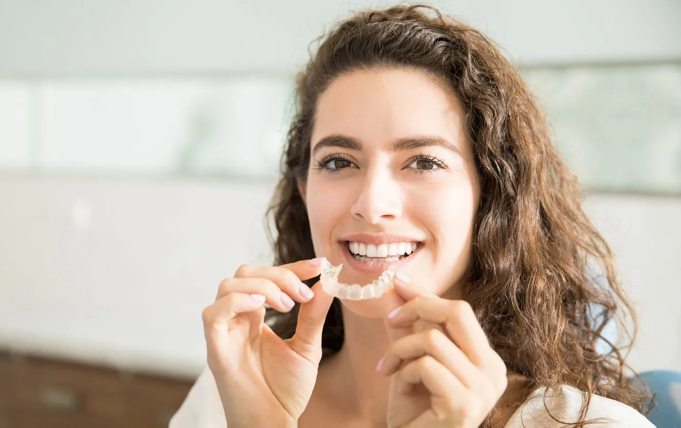 Young woman holding clear aligners and smiling at the camera during a consultation, representing the use of custom-fit clear aligner therapy for discreet orthodontic correction