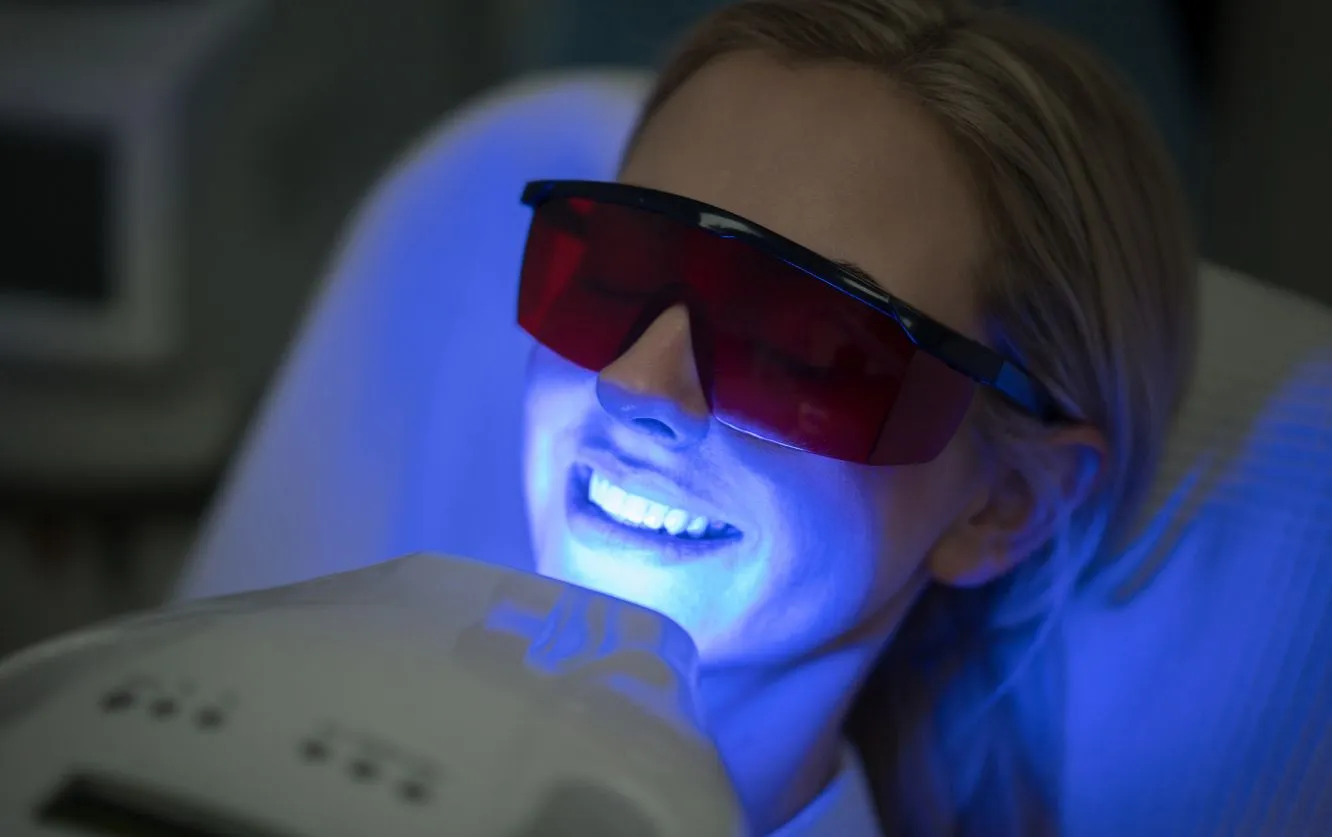 Female patient receiving professional teeth whitening under a blue light device while reclining in a treatment chair, emphasizing non-invasive cosmetic enhancement