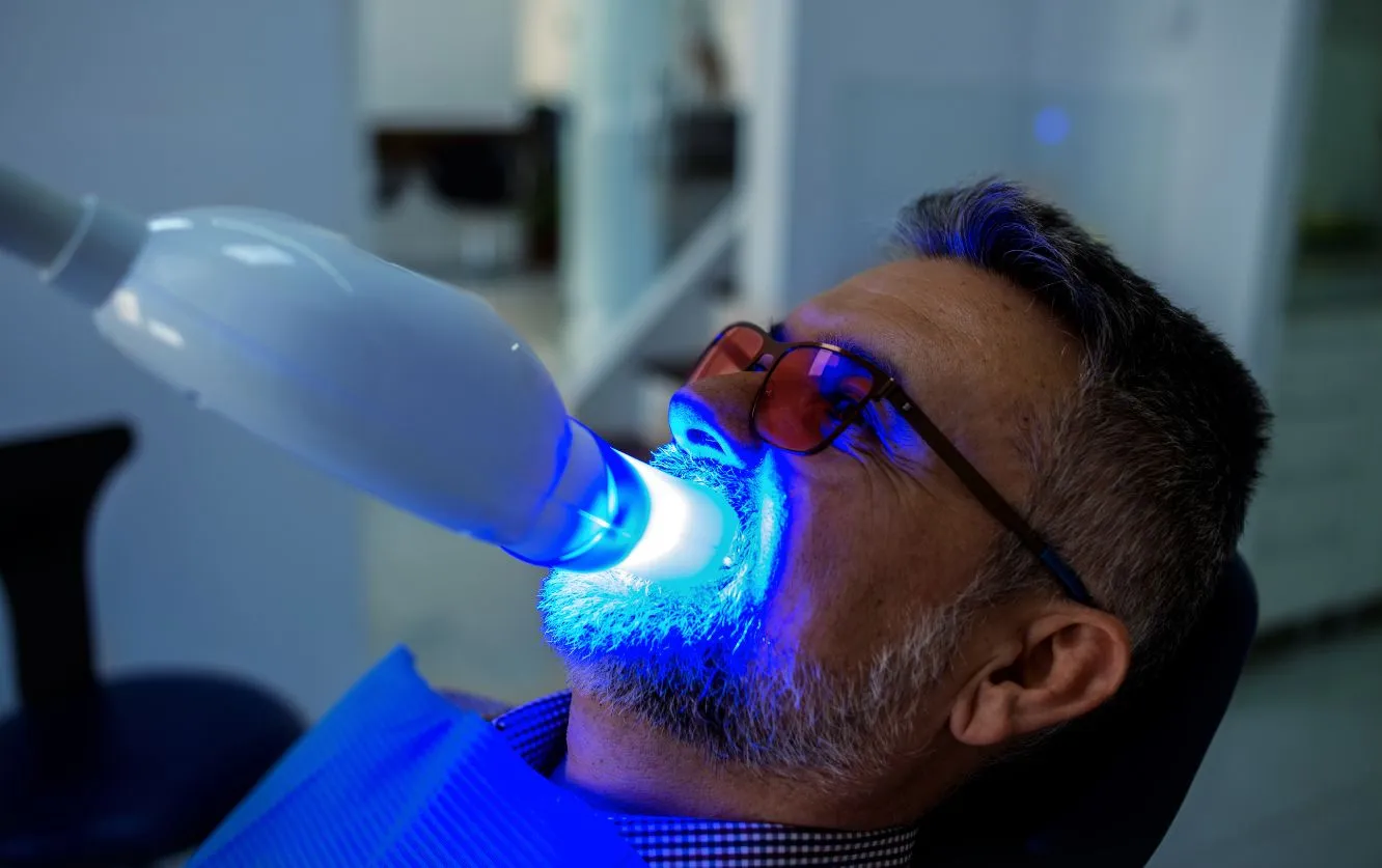 Male patient undergoing in-office teeth whitening procedure using a blue LED curing light while reclined in a dental chair, highlighting advanced cosmetic treatment options