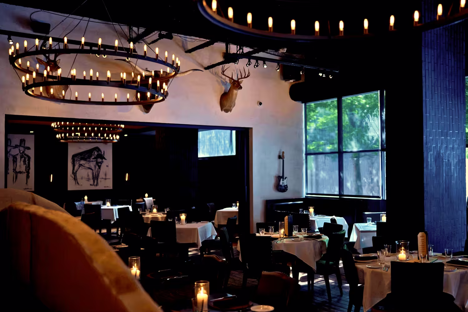 Dimly lit dining room at Lonesome Dove Austin with white tablecloths, lit candles, mounted deer head, and circular chandeliers.