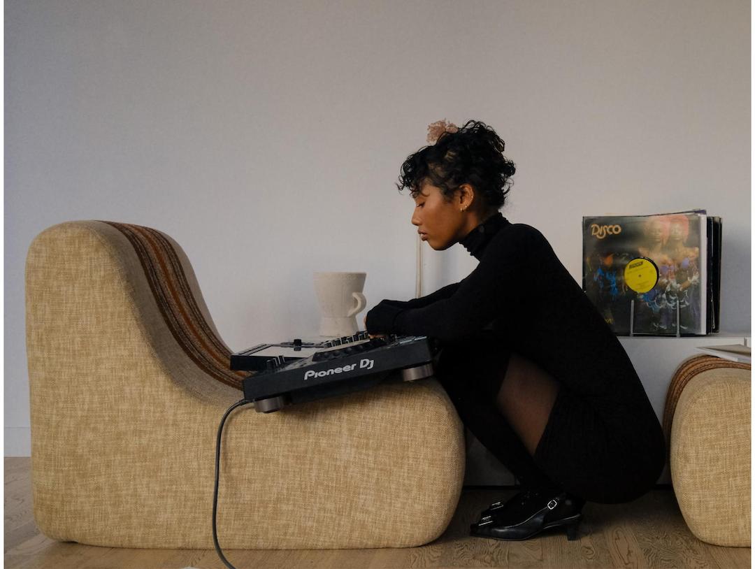Minzi Roberta crouches down at the foot of a chair, with her mixer propped up on it. She is dressed in all black for this artist portrait.