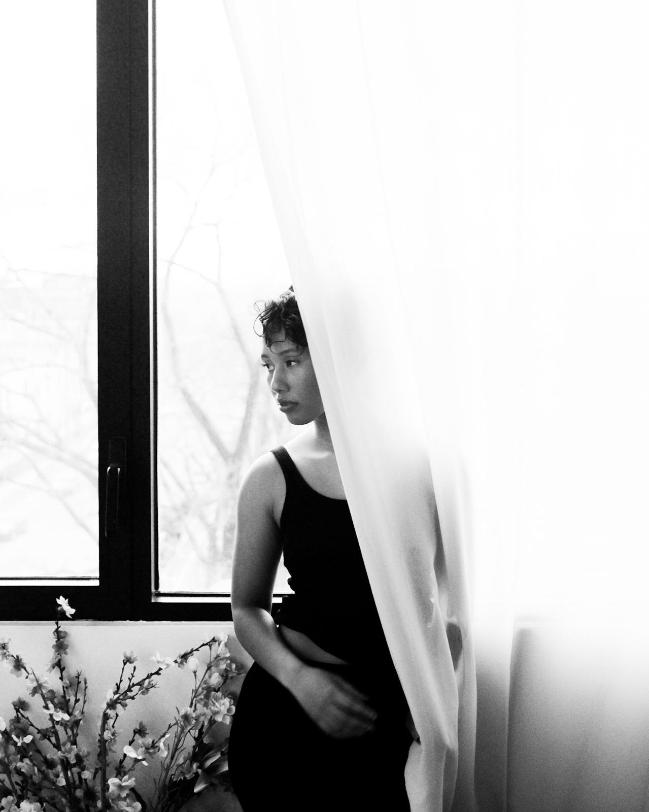 Minzi Artist Asset, she stands behind a light soft curtain by a window in black & white