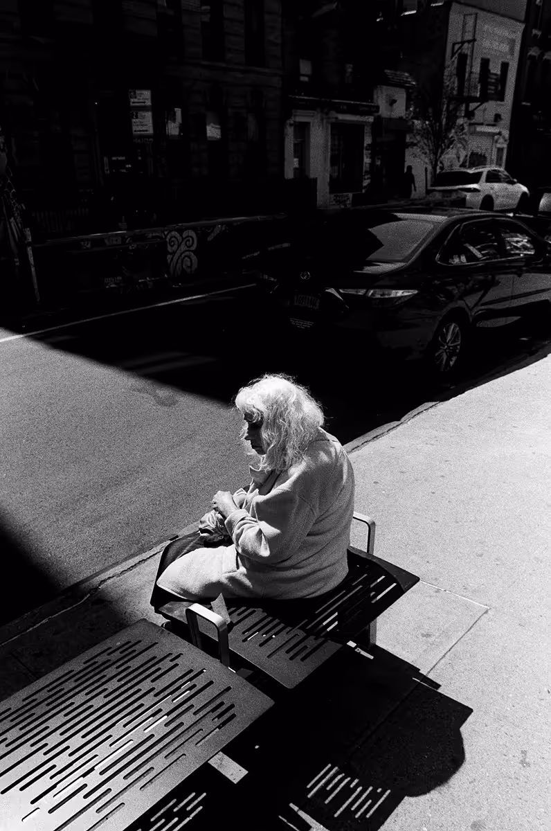 Elderly woman with white hair sitting on sunny sidewalk bench