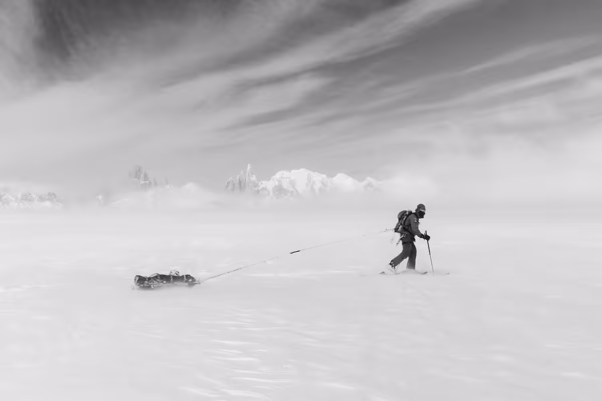 Skier pulling sled across windswept glacier with mountain peaks emerging from clouds